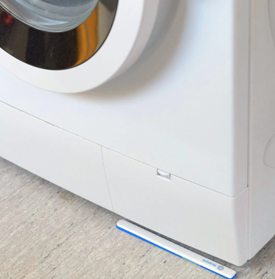 A Strips MS +Drip sensor lying flat on the floor in the gap beneath a washing machine, where a leak would first collect