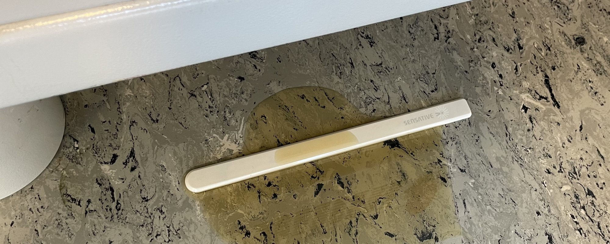 Ein Strips-MS-+Oil-Sensor auf einem Werkstattboden in einer sich ausbreitenden Lache Hydrauliköl, das Öl sichtbar in das Absorberpad entlang eines Teils des Streifens gezogen